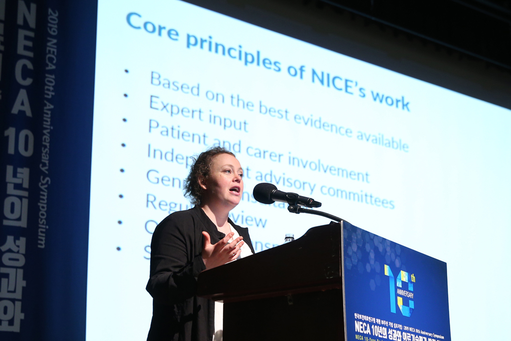 NICE's Dr Grace Jennings on stage at NECA's 10th anniversary symposium, with a presentation slide titled 'Core principles of NICE's work' on a screen in the background