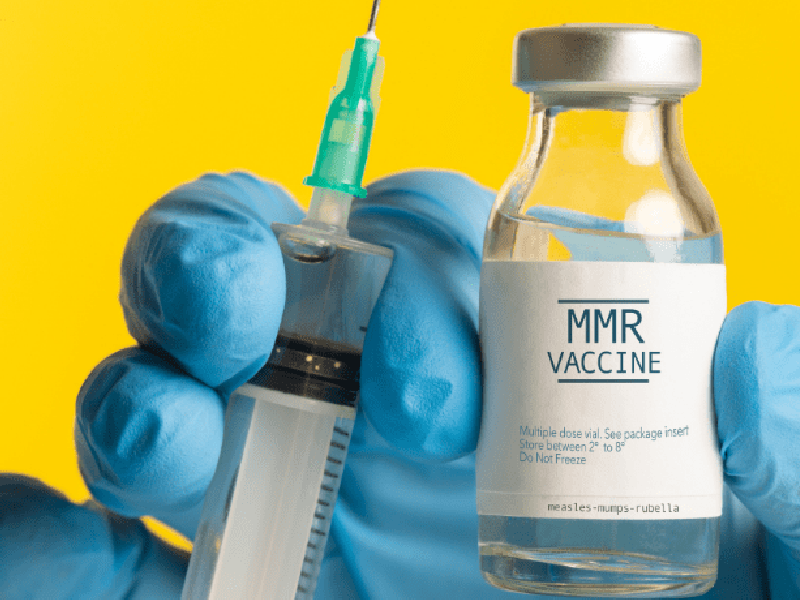 A hand wearing a medical glove holding a vial of MMR vaccine and a syringe