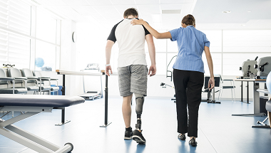 A physical therapist supports a man with a prosthetic leg as he walks