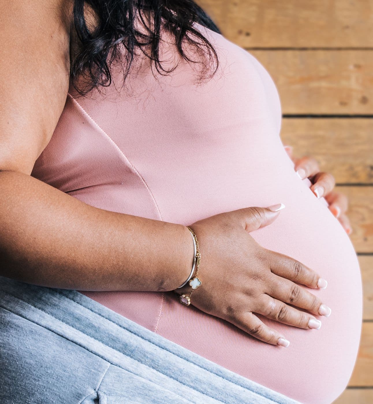 A pregnant woman wearing a pink top gently cradles her belly with both hands, standing against a wooden wall.
