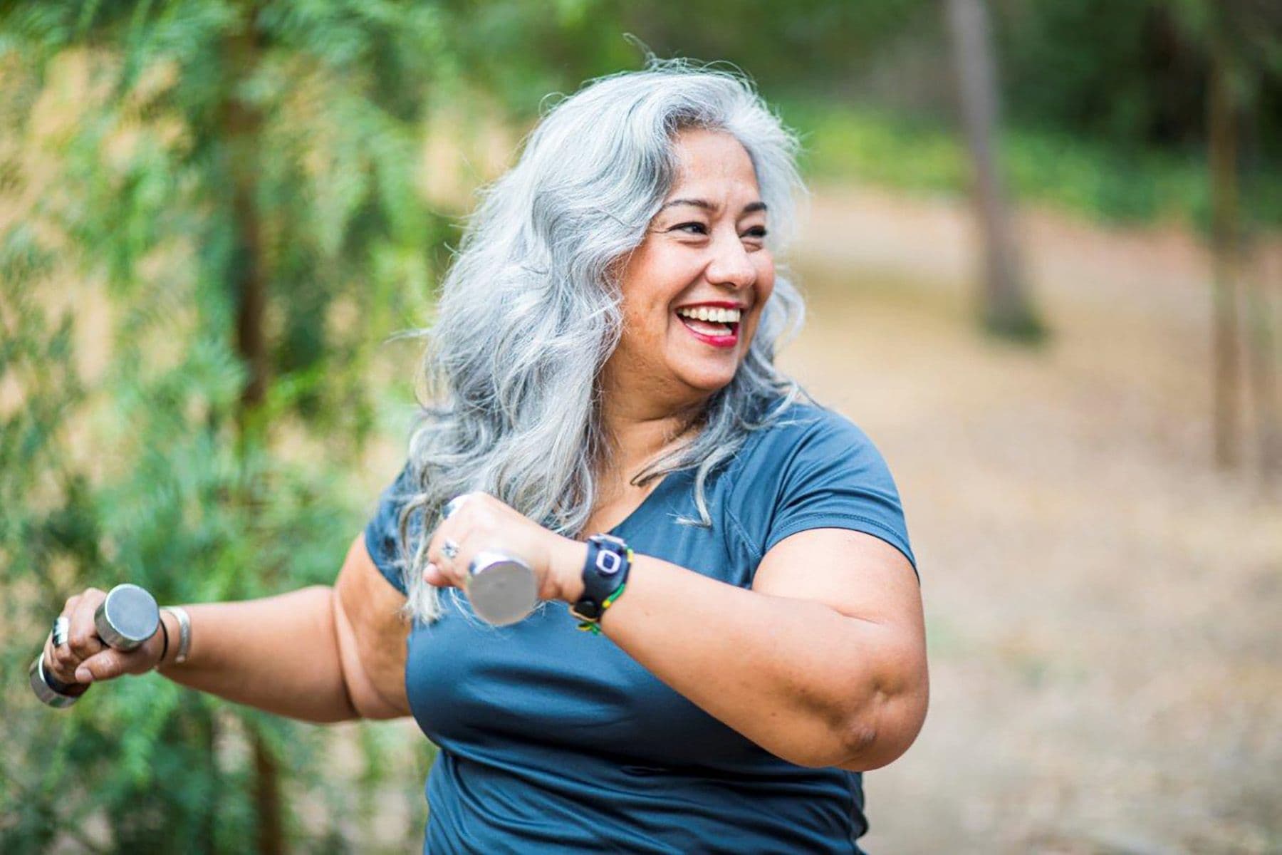 A smiling woman with gray hair exercises with dumbbells outdoors, wearing a blue shirt and a watch, with blurred greenery in the background.