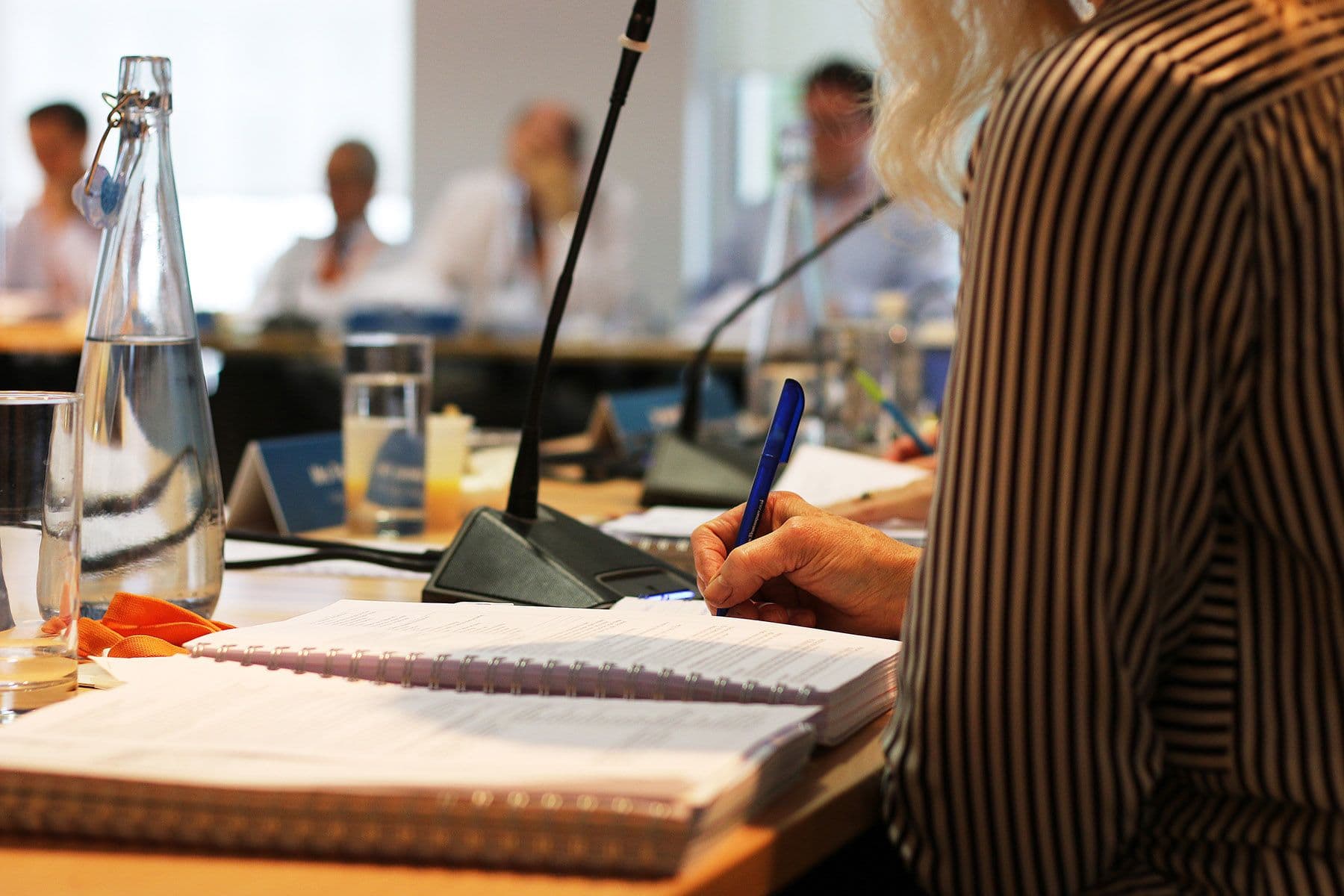 A person in a striped shirt takes notes in a notebook during a meeting. A microphone and a bottle of water are on the table.
