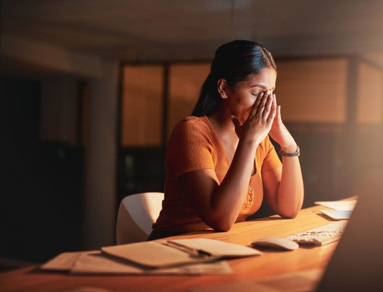 A woman sitting at a desk with her hands on her face, experiencing a migraine
