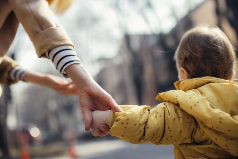 Adult holding a child's hand on a street. The child is wearing a yellow coat, and the scene evokes warmth and guidance.