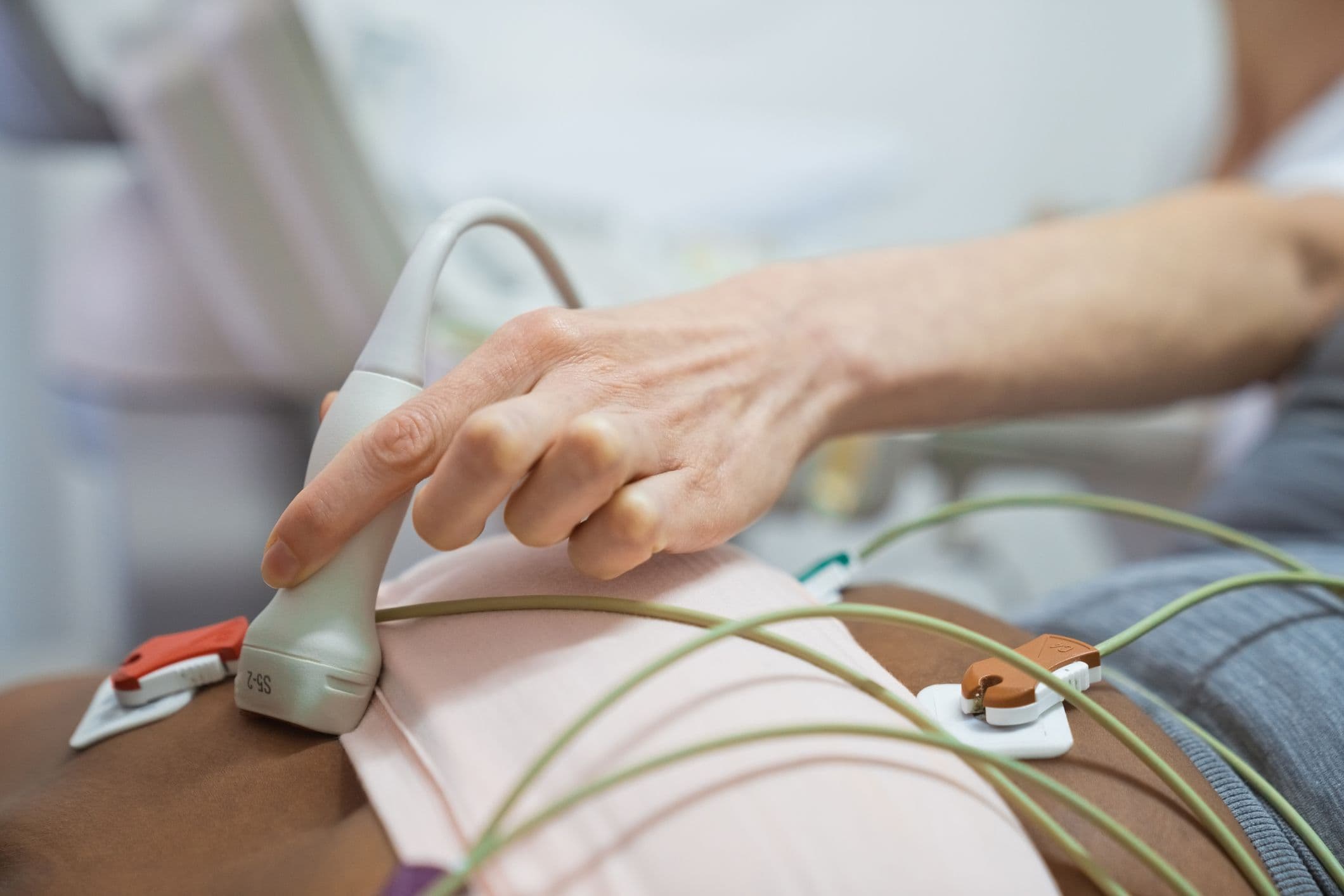 Close-up of a hand holding an ultrasound probe on a person's abdomen, connected to monitoring electrodes with wires.