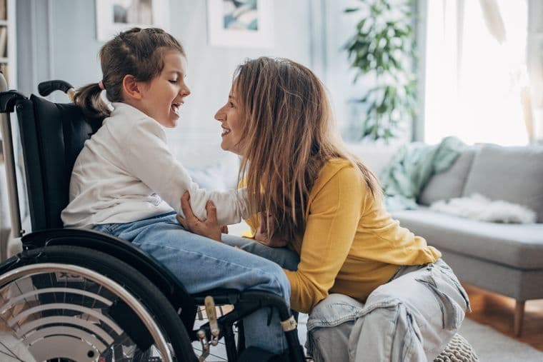 A woman in a yellow sweater smiles warmly at a young child in a wheelchair, sharing a joyful moment in a cozy living room.