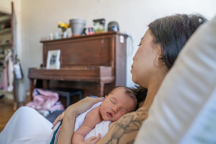 A new Mother lies on cushions while holding her infant to her chest with a piano and ornaments in the background.