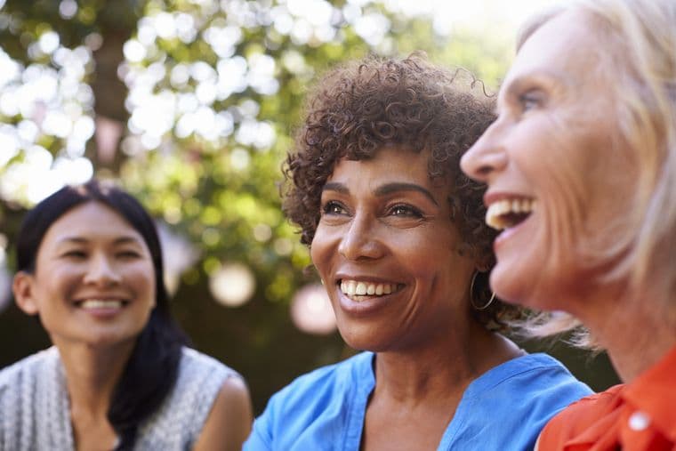 Three women smiling and laughing outdoors, with sunlight filtering through the trees in the background.