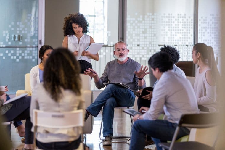 A researcher standing in a room with a group of people sat on chairs in a circle talking