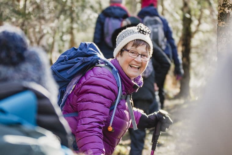 A woman in a purple jacket and knit hat smiles while hiking in a forest with a group, holding trekking poles and wearing a backpack.