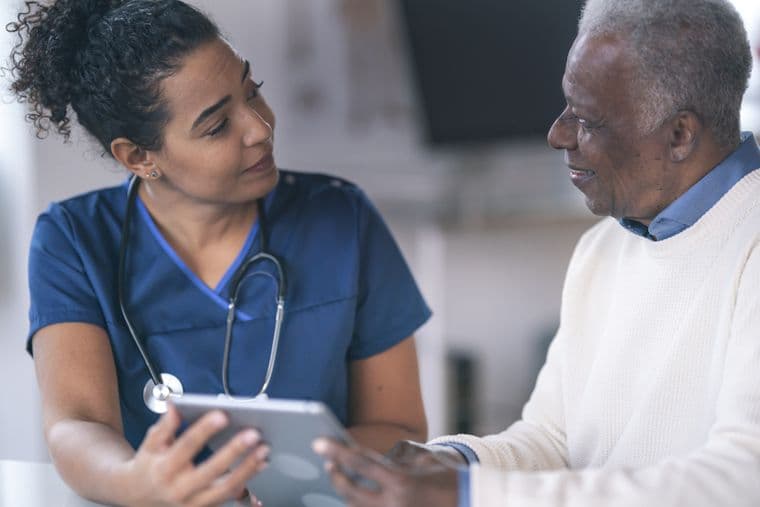 A mixed-race female doctor is meeting with a patient. The patient is a senior black man. The two individuals are seated next to each other at a table in a medical examination room. The medical professional is holding a wireless tablet computer. The two individuals are looking at each other. They are discussing the patient's test results and medical history.