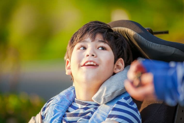 A smiling child in a wheelchair, wearing a striped shirt and blue jacket, looks upward outdoors with a blurred green background.