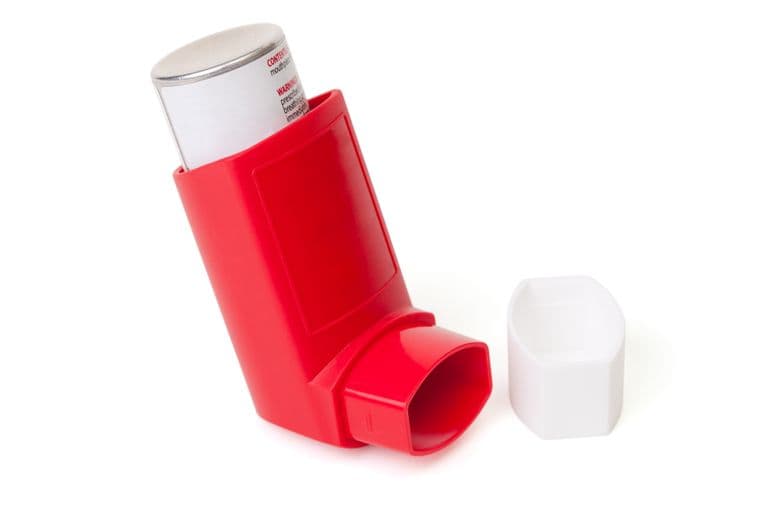 A red asthma inhaler with a silver canister, lying on its side next to a white cap, on a white background.