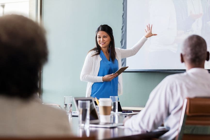 While gesturing toward a projected image, a medical technology developer discusses a new technology. She is holding a digital tablet
