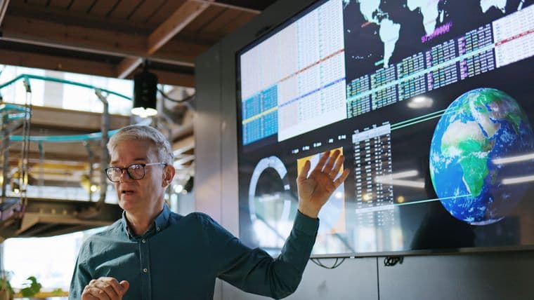 a mature man conducting a seminar with the aid of a large screen displaying graphs and images of the earth.