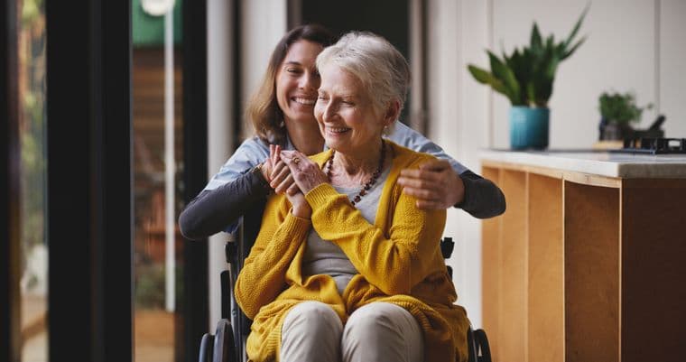 A caregiver embraces a smiling elderly woman in a wheelchair, creating a warm and supportive atmosphere in a bright room with plants.