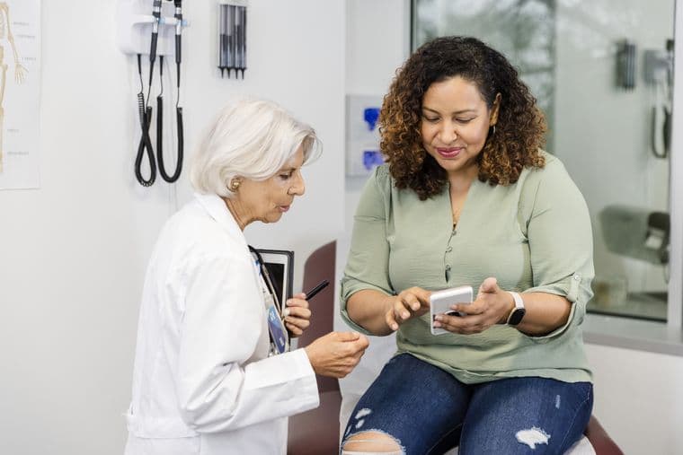 A doctor and a patient sit in a medical office. The patient, holding a smartphone, listens attentively as the doctor explains something.