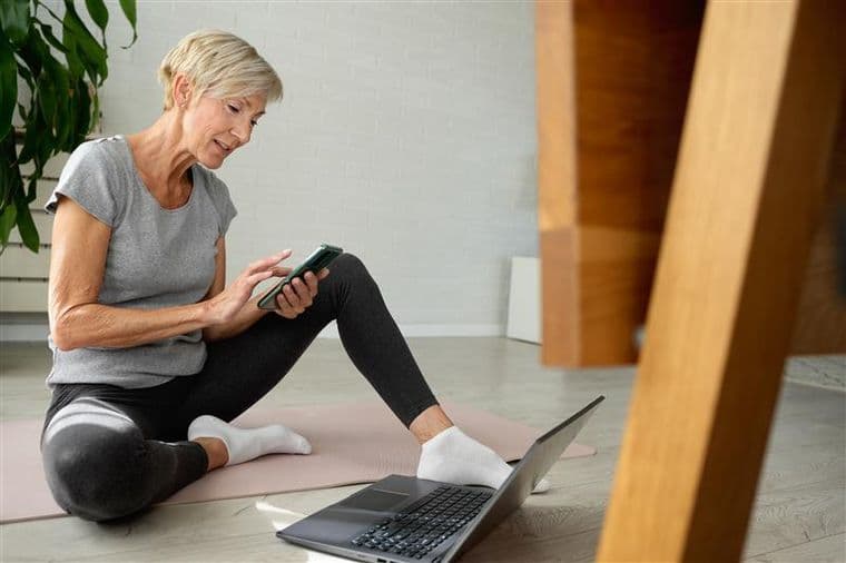 Older woman in workout clothes sits on a yoga mat, holding a phone, with a laptop in front of her. A large plant is in the background.