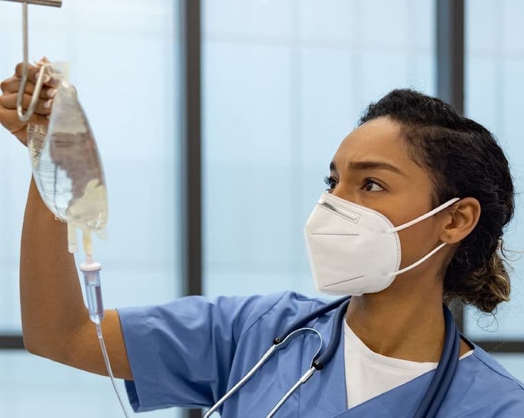 A nurse in blue scrubs and a mask examines an IV drip in a hospital setting